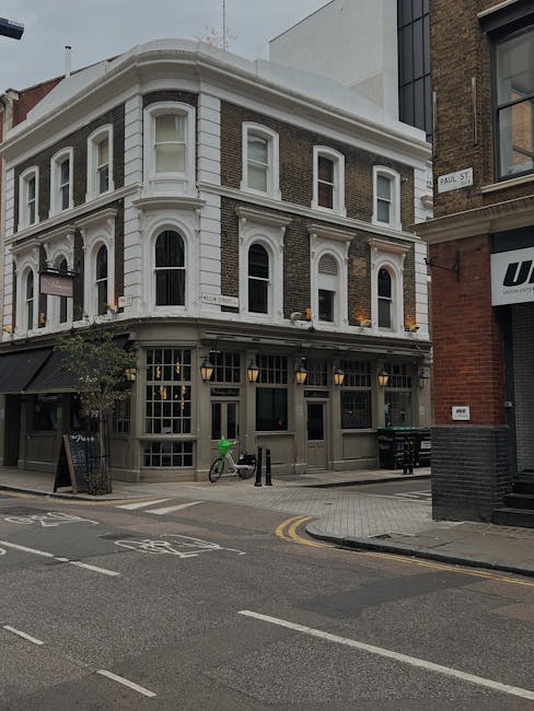 A three-story corner building on Brixton Road features large front windows on the ground floor and smaller arched windows on the upper floors, with a white and dark brick exterior. In front of the building, a man with Van Brixton, a professional removals service, is loading a vehicle with various household items, including cardboard boxes wrapped in plastic, using a trolley and lifting equipment. The loading area is on the paved street level with a bicycle secured to a bike rack near the entrance. The scene captures the process of home relocation, with furniture transport preparations evident at the street-facing side of the property, under daylight with overcast weather conditions.