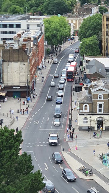Aerial view of a busy urban street near Brixton tube station, showing a line of vehicles including cars and a double-decker bus traveling along a curved road. On the left side, there are multi-storey buildings with storefronts and pedestrians walking on the sidewalk. To the right, there are smaller residential buildings and shops, with bicycles parked on the pavement. Several large green trees line the street, providing shade and greenery in the city environment. The scene suggests daytime with natural light illuminating the street scene, indicative of active city life suitable for house removals or moving services. The image captures the typical traffic flow and urban landscape that Man with Van Brixton might encounter during property relocation or furniture transport processes in the area.