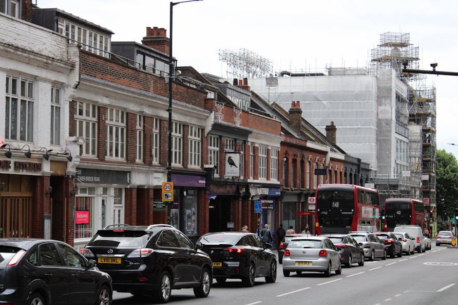A three-story corner building on Brixton Road features large front windows on the ground floor and smaller arched windows on the upper floors, with a white and dark brick exterior. In front of the building, a man with Van Brixton, a professional removals service, is loading a vehicle with various household items, including cardboard boxes wrapped in plastic, using a trolley and lifting equipment. The loading area is on the paved street level with a bicycle secured to a bike rack near the entrance. The scene captures the process of home relocation, with furniture transport preparations evident at the street-facing side of the property, under daylight with overcast weather conditions.